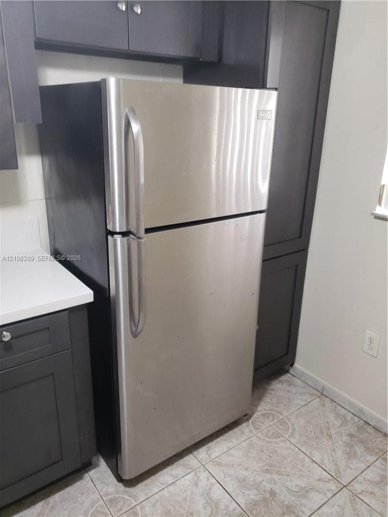 Stainless steel top-freezer refrigerator, dark-finish shaker cabinetry, light-toned stone-finish countertops, and ceramic tile flooring