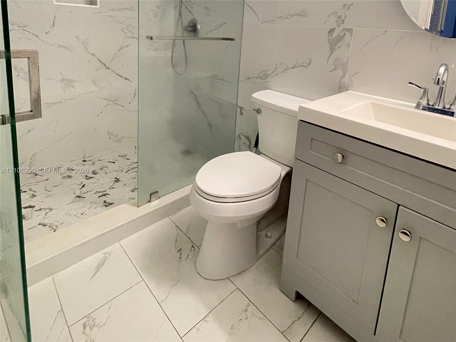 Bathroom featuring a walk-in glass-enclosed shower with tiled surround, stone-patterned floor tile, grey shaker-style vanity cabinet, and rectangular sink