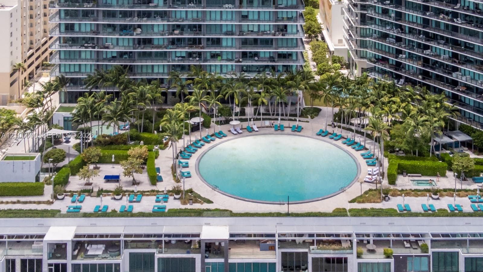 Circular swimming pool surrounded by paved decking, manicured landscaping, mature palm trees, and an integrated hot tub