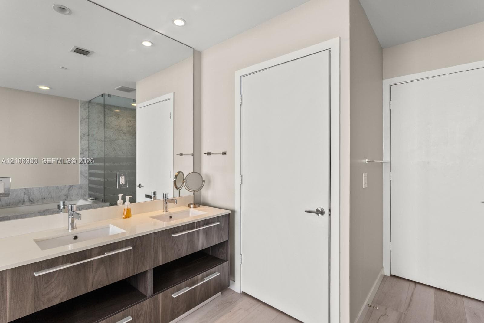 Double vanity featuring a light-toned countertop with integrated dual sinks, wood-finish cabinetry with linear hardware, full-width wall mirror, and recessed lighting