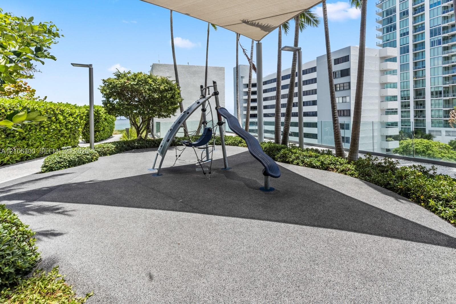 Outdoor recreation area featuring a play structure with a slide, rubberized surfacing, overhead shade canopy, perimeter landscaping, and vertical lighting fixtures