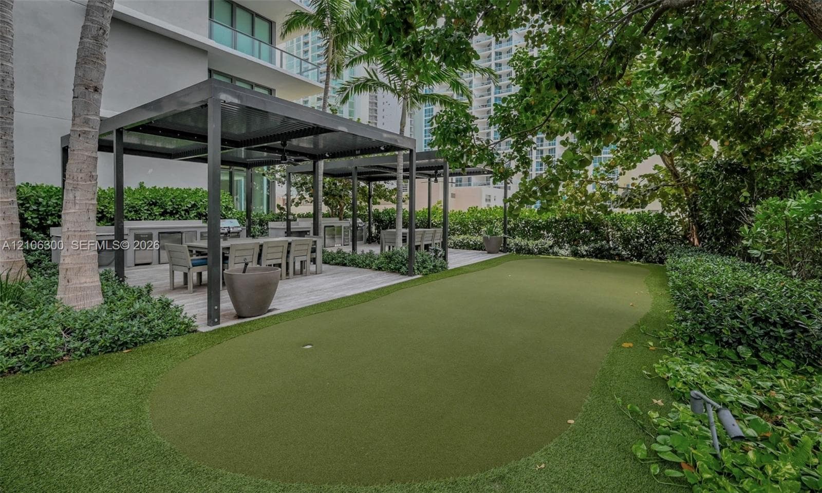 Outdoor terrace featuring a synthetic turf putting green, dual metal-framed pergolas, a built-in outdoor kitchen, and mature privacy hedges