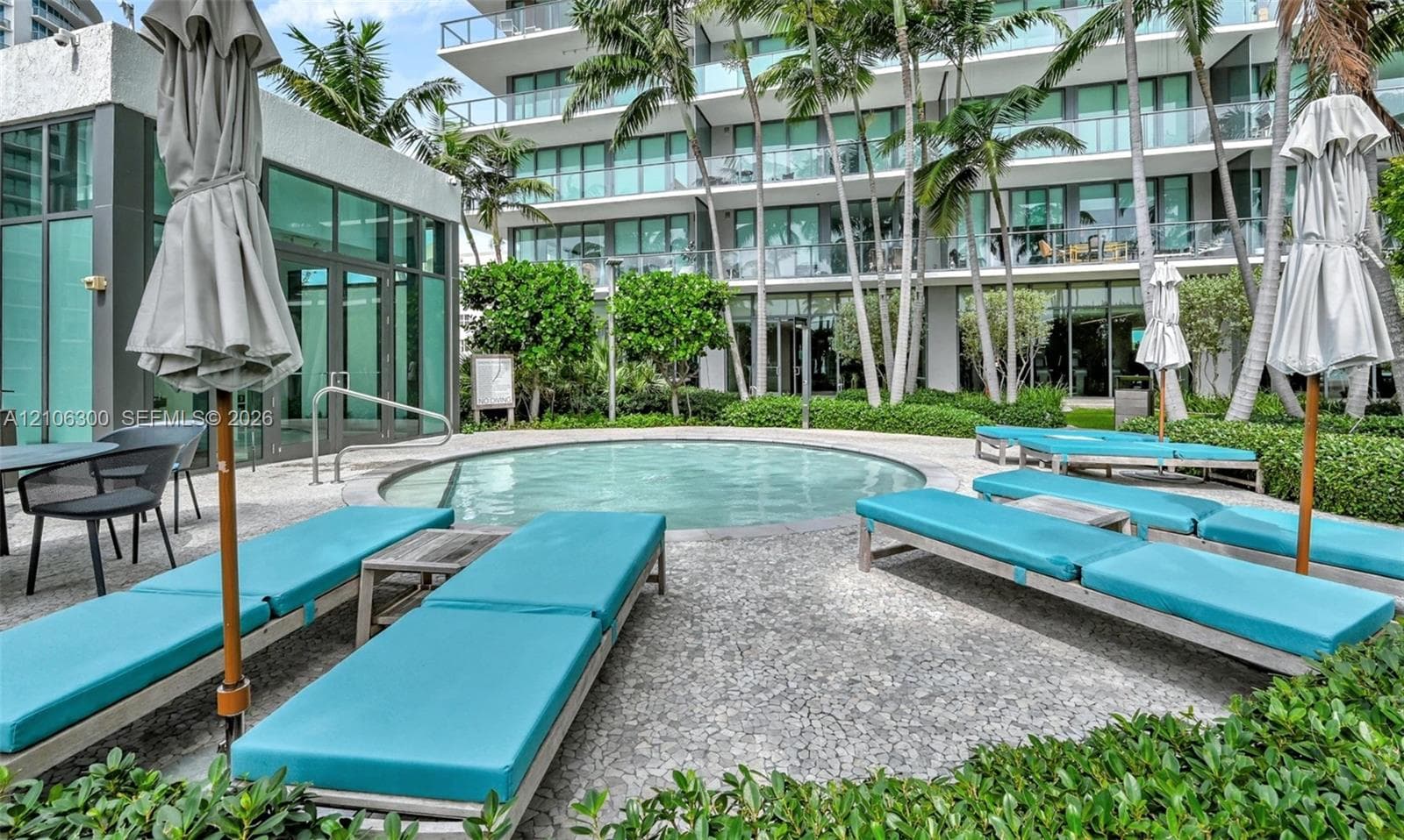 Circular swimming pool surrounded by a stone-paved deck, landscaped garden beds, and palm trees
