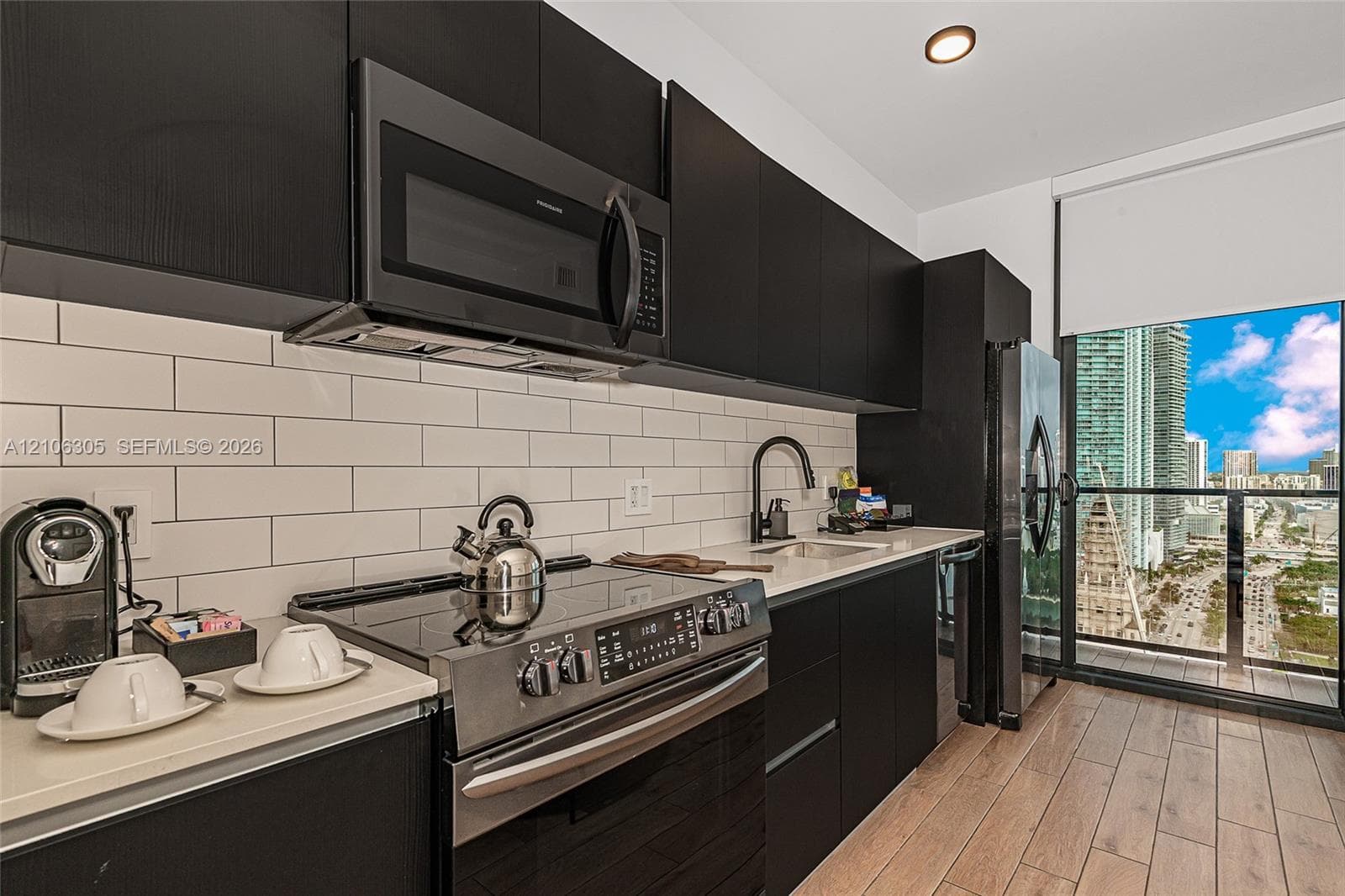 Modern kitchen featuring black cabinetry, light-toned stone-finish countertops, white subway tile backsplash, stainless steel appliances, and wood-finish flooring