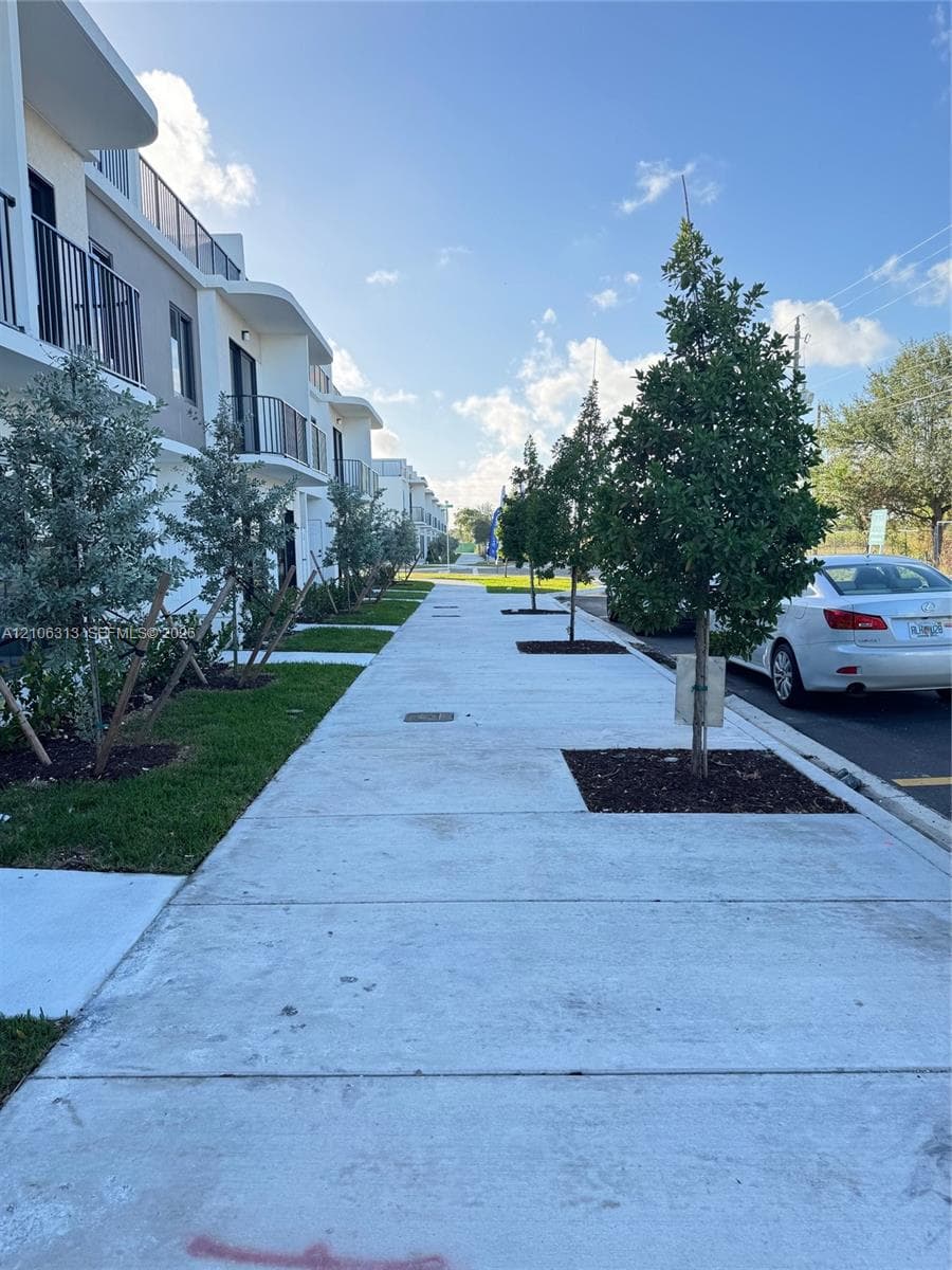 Multi-story exterior featuring a modern facade with rounded architectural accents, integrated metal balcony railings, concrete sidewalk, and landscaped tree-lined beds