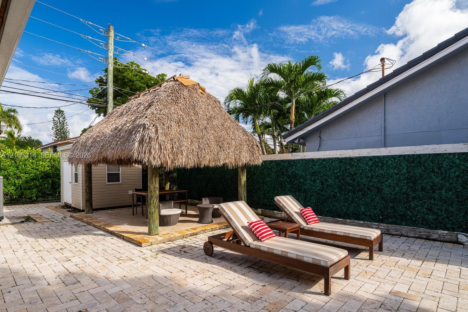 Outdoor patio featuring interlocking stone pavers, a thatched-roof tiki hut, and privacy hedging
