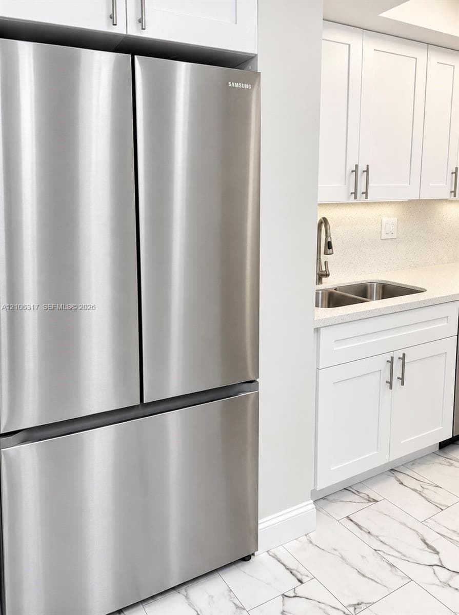 Stainless steel French door refrigerator, white shaker-style cabinetry with brushed metal hardware, solid-surface countertops, dual-basin stainless steel sink with high-arch faucet, and patterned tile flooring