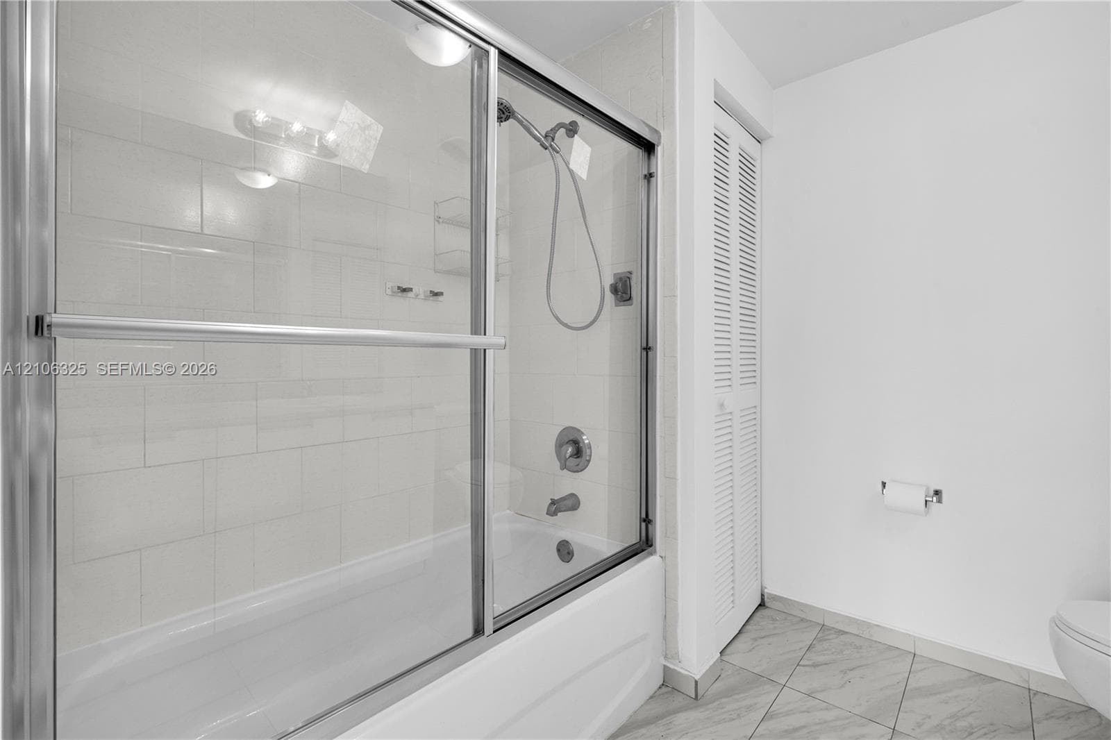 Bathroom featuring a tub-shower combination with a sliding glass enclosure, white tile wall surround, chrome shower fixtures, and neutral floor tile