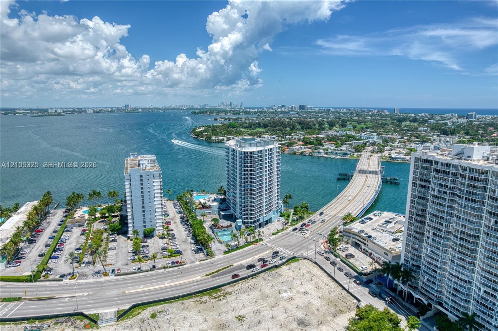 Aerial perspective featuring extensive water frontage, multiple high-rise structures, paved parking facilities, mature palm landscaping, and an expansive bridge crossing