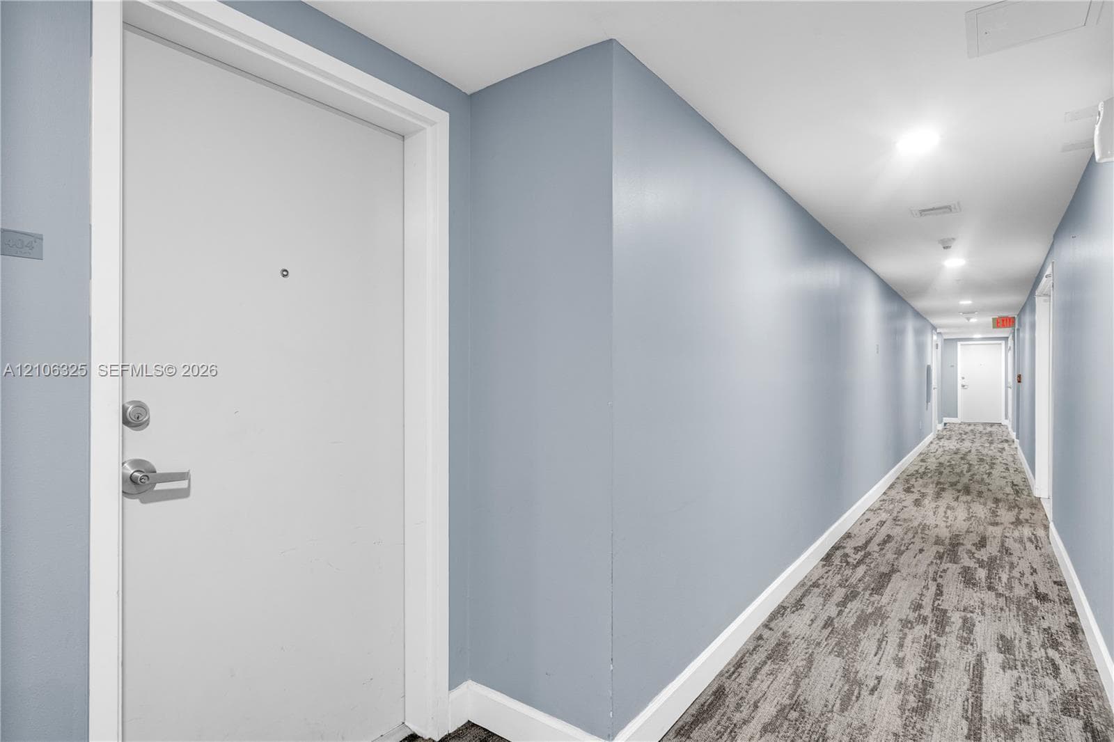 Hallway featuring light blue walls, neutral-toned patterned carpeting, recessed ceiling lighting, and a solid-core door equipped with a chrome lever handle and deadbolt