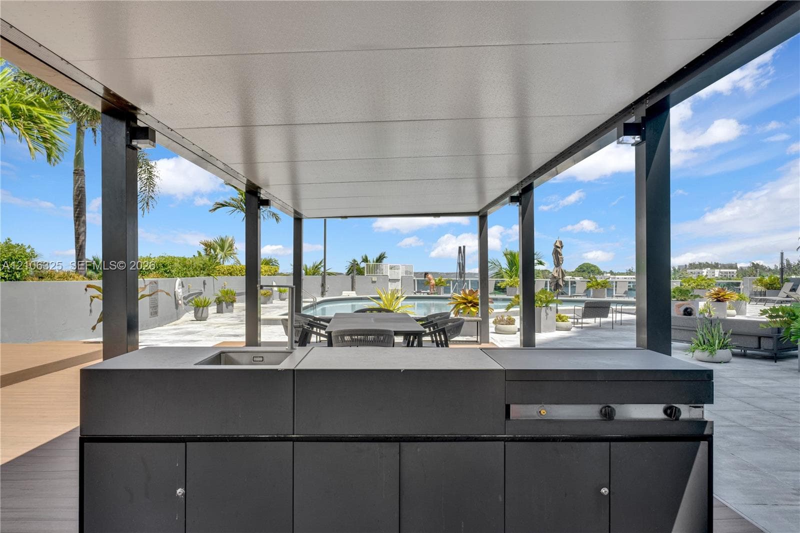 Covered outdoor kitchen featuring a flat-top grill, integrated sink, and modern cabinetry, situated on a tiled terrace with a swimming pool