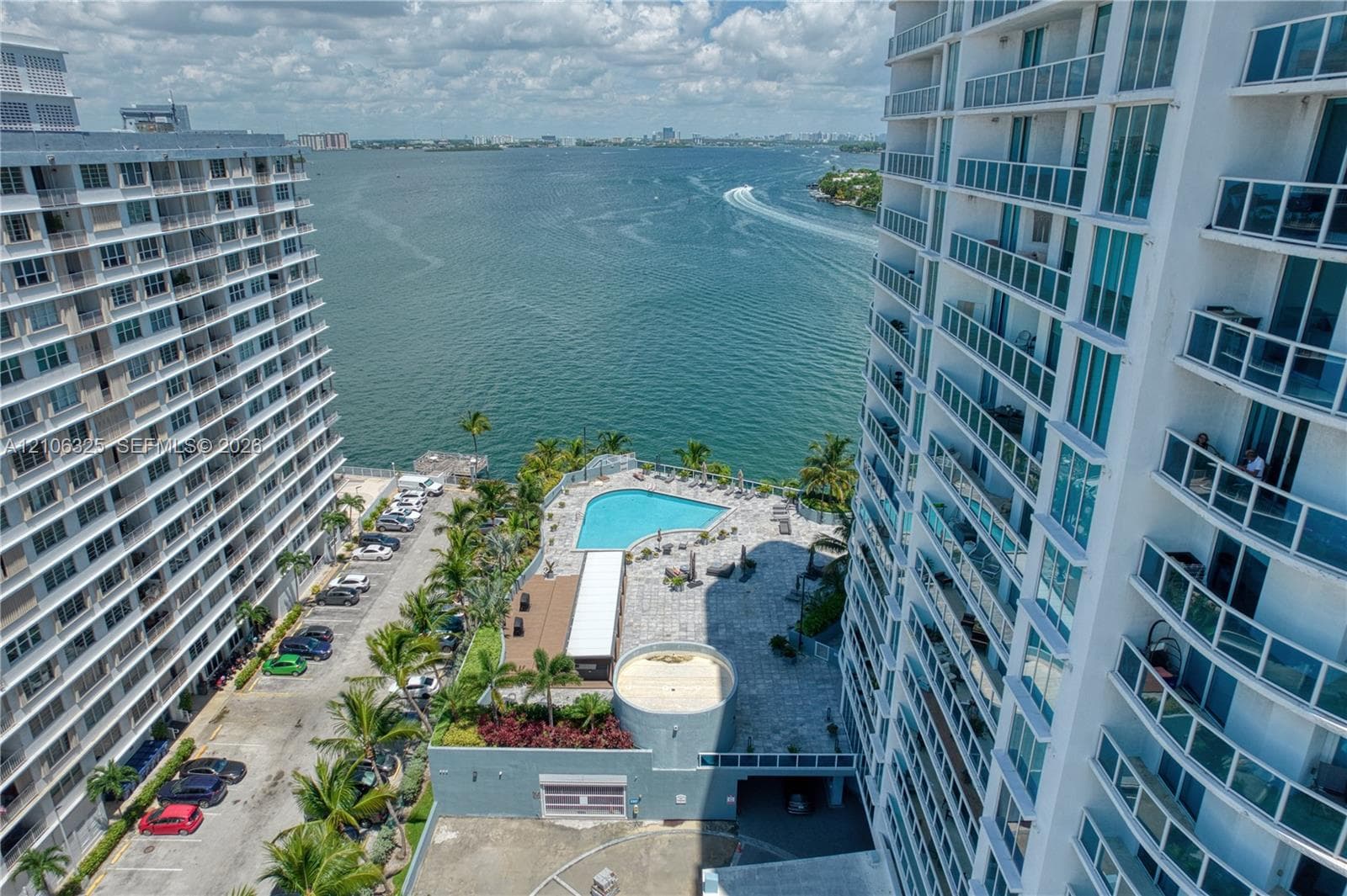High-rise residential exterior featuring private balconies with glass railings, an outdoor swimming pool, paved sundeck, and palm tree landscaping overlooking the water