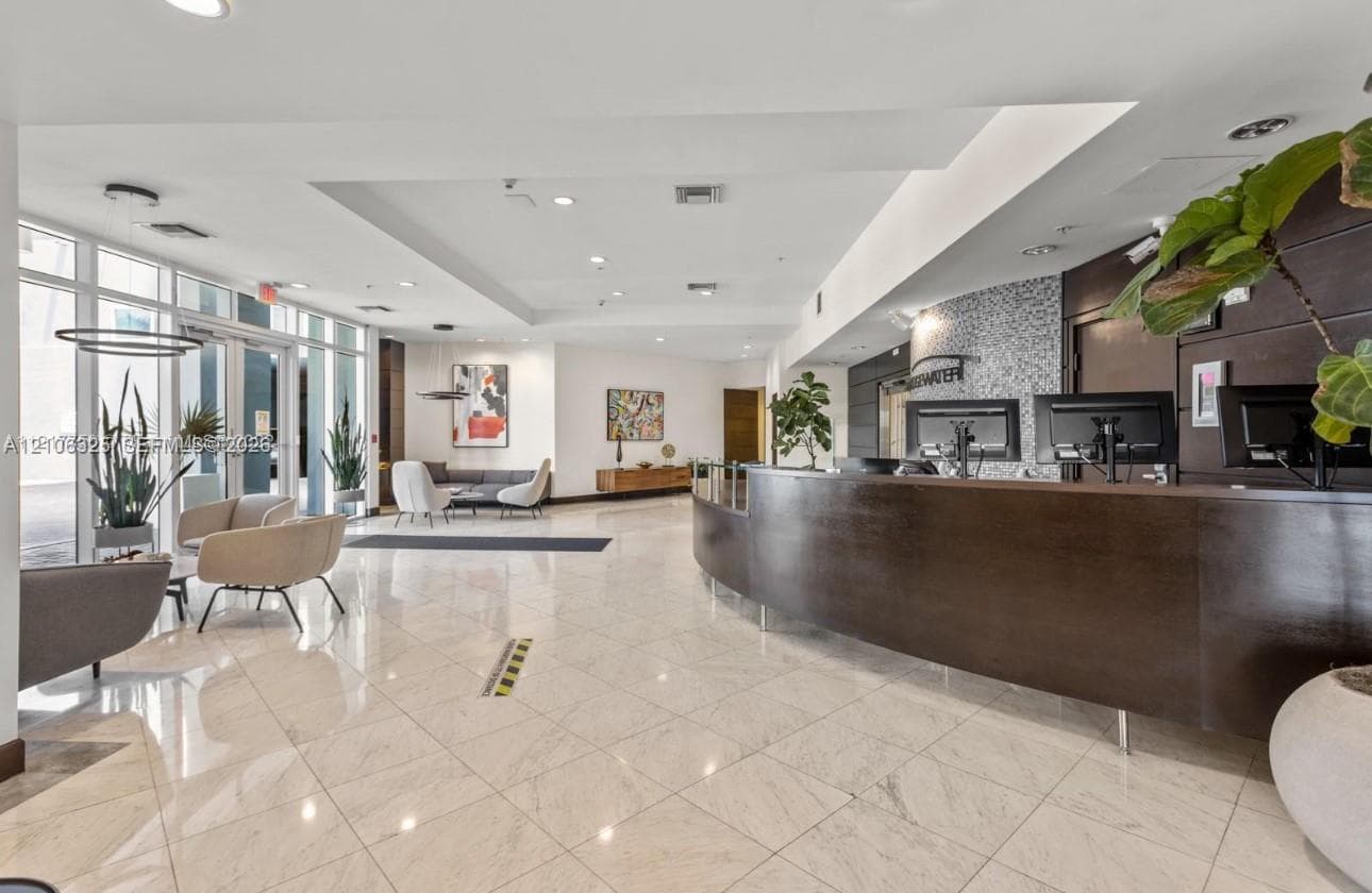 Spacious lobby featuring polished stone flooring, a curved wood-finish reception desk, recessed ceiling lighting, and floor-to-ceiling glass windows