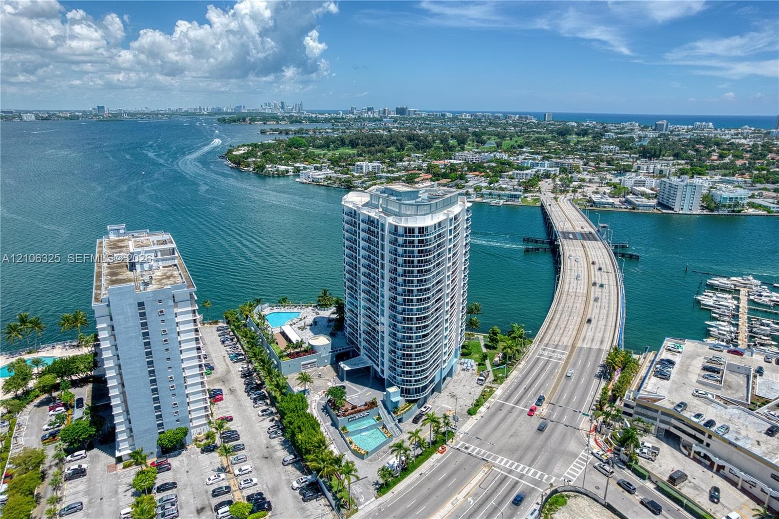 High-rise condominium tower situated on a waterfront peninsula, featuring exterior balconies, a ground-level swimming pool, and dedicated surface parking