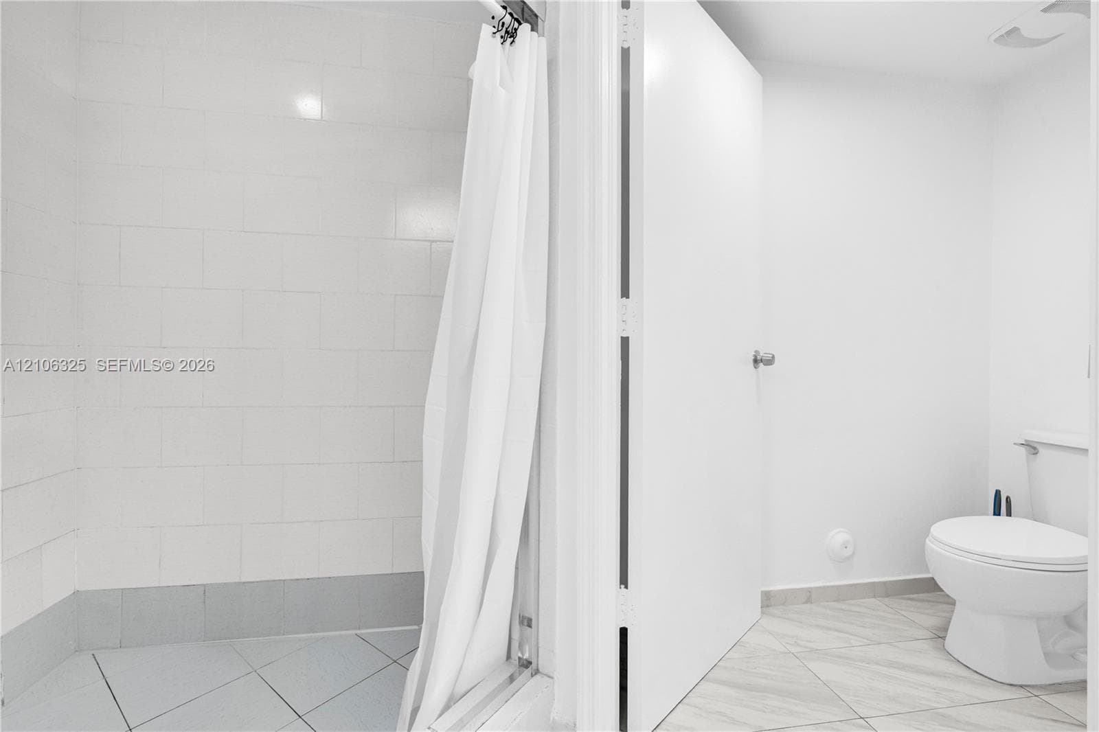 Bathroom featuring a shower stall with white tile surround, light-toned stone-finish floor tile, and a standard porcelain toilet