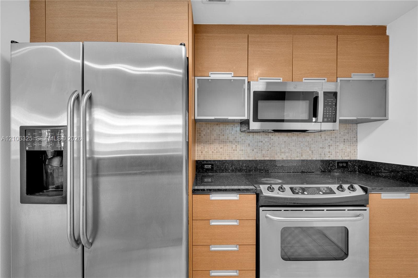 Kitchen featuring flat-panel wood-finish cabinetry, stainless steel appliances, dark stone countertops, mosaic tile backsplash, and frosted glass cabinet accents