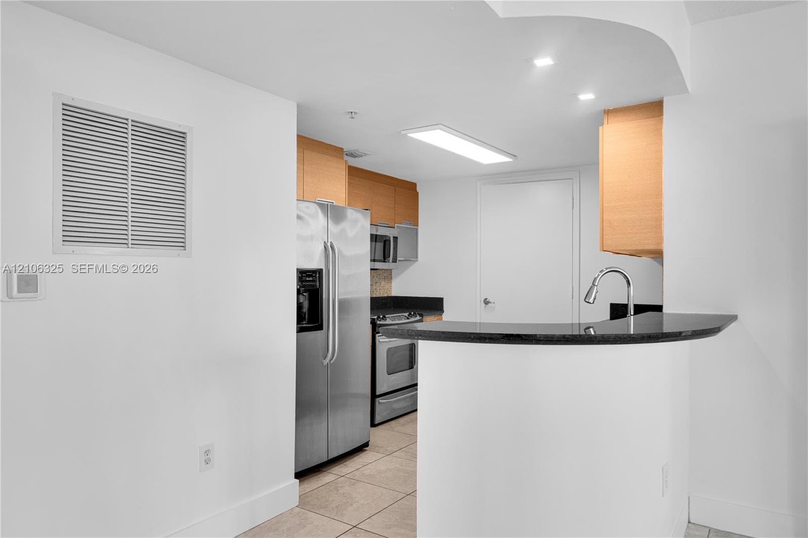 Kitchen featuring natural wood-finish cabinetry, stone-surfaced countertops, a curved breakfast bar, stainless steel appliances, and tile flooring