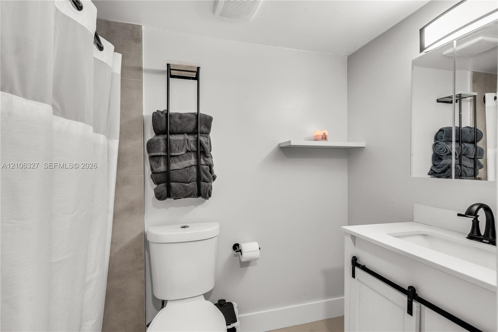 Bathroom featuring a white vanity with a composite countertop, matte black faucet, mirrored medicine cabinet, wall-mounted shelving, and a tiled shower surround