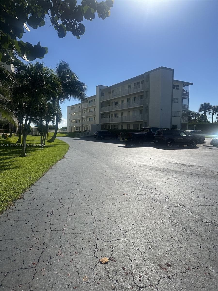 Multi-story residential building featuring covered balconies, asphalt parking area, and manicured green space with mature palms