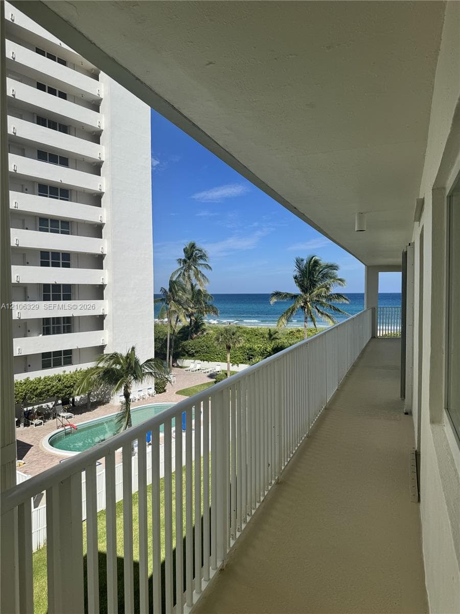 Covered balcony featuring a white metal railing, concrete flooring, ocean horizon, and direct oversight of the swimming pool and palm landscaping