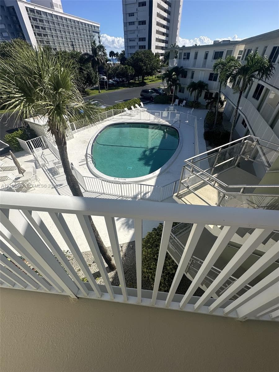 Elevated balcony with a white metal railing overlooking a community pool area with a paved deck, perimeter fencing, and mature palm trees