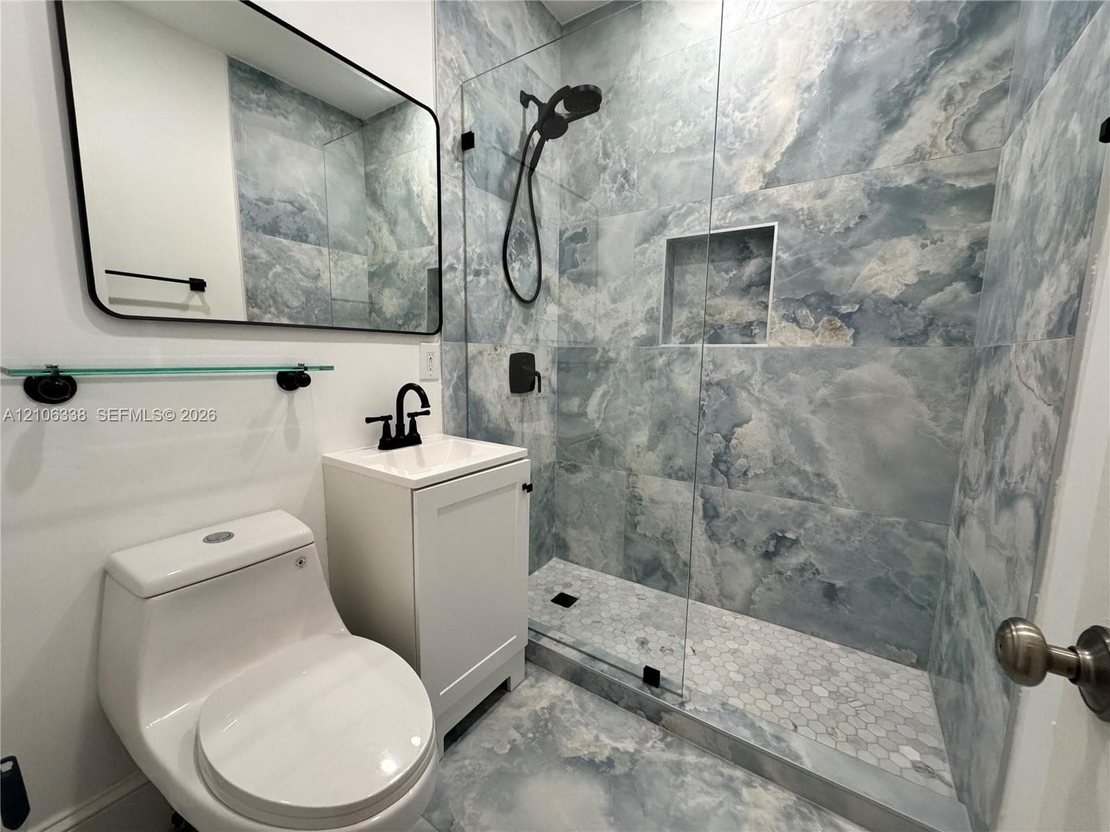 First Floor. Walk-in shower featuring large-format patterned stone-finish tile, glass enclosure, and built-in wall niche, paired with a white vanity and matte black hardware