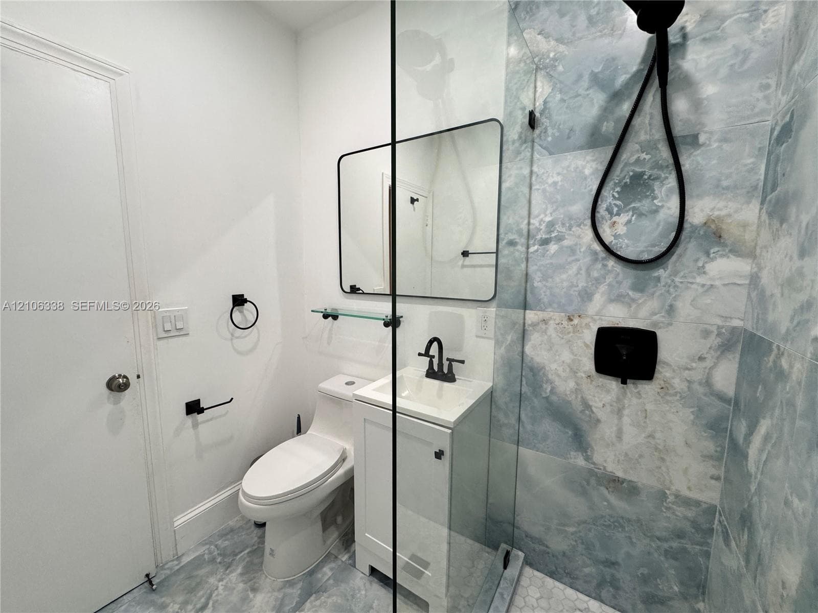 Walk-in shower with floor-to-ceiling stone-finish tile, glass partition, vanity with integrated basin, matte black hardware, and rectangular wall-mounted mirror