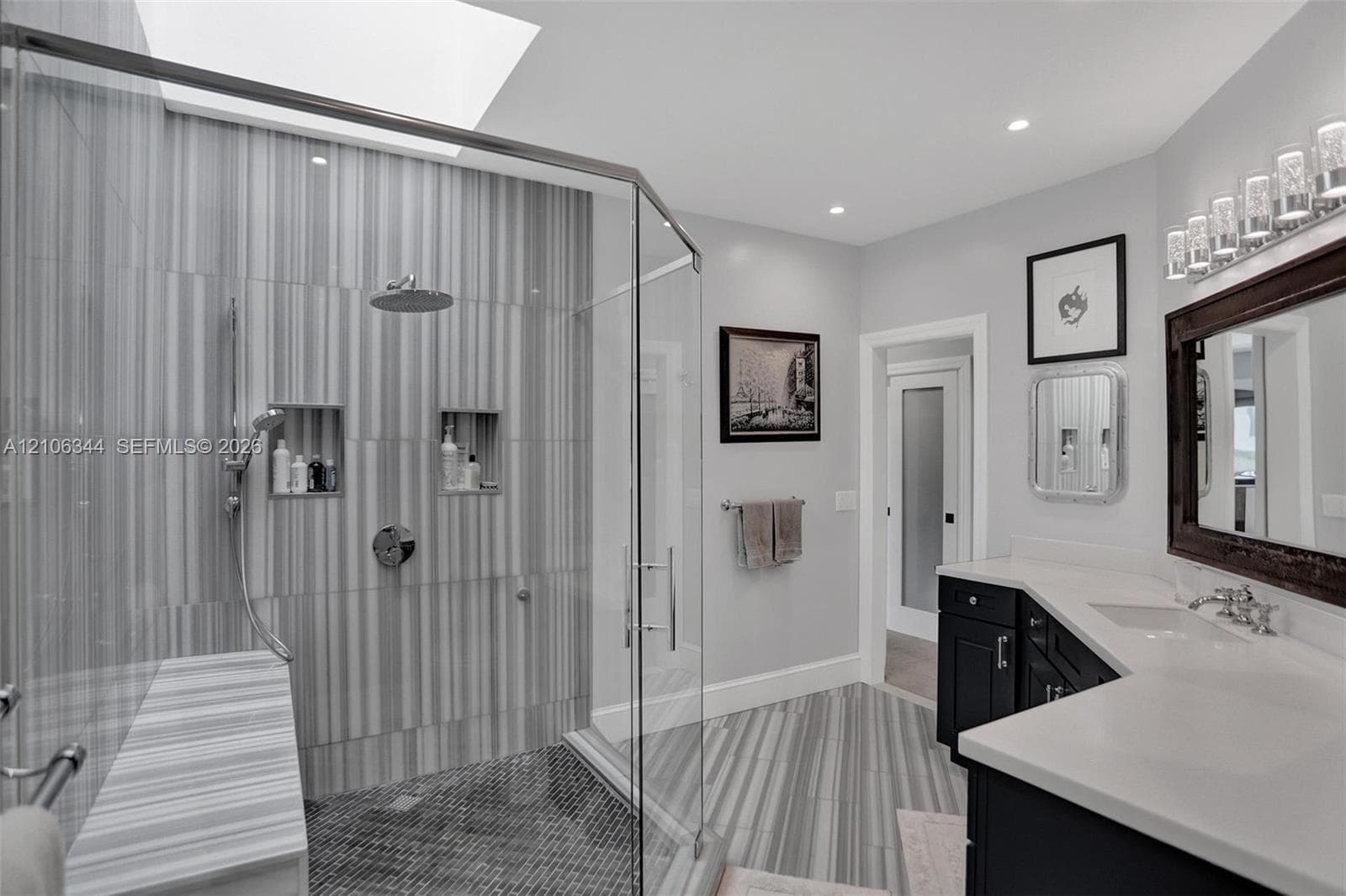 Walk-in shower with vertical tile surround, dual recessed shower niches, overhead rainfall showerhead, glass enclosure, and stone-finish bench seat, accompanied by a dual-sink vanity with solid-surface countertops