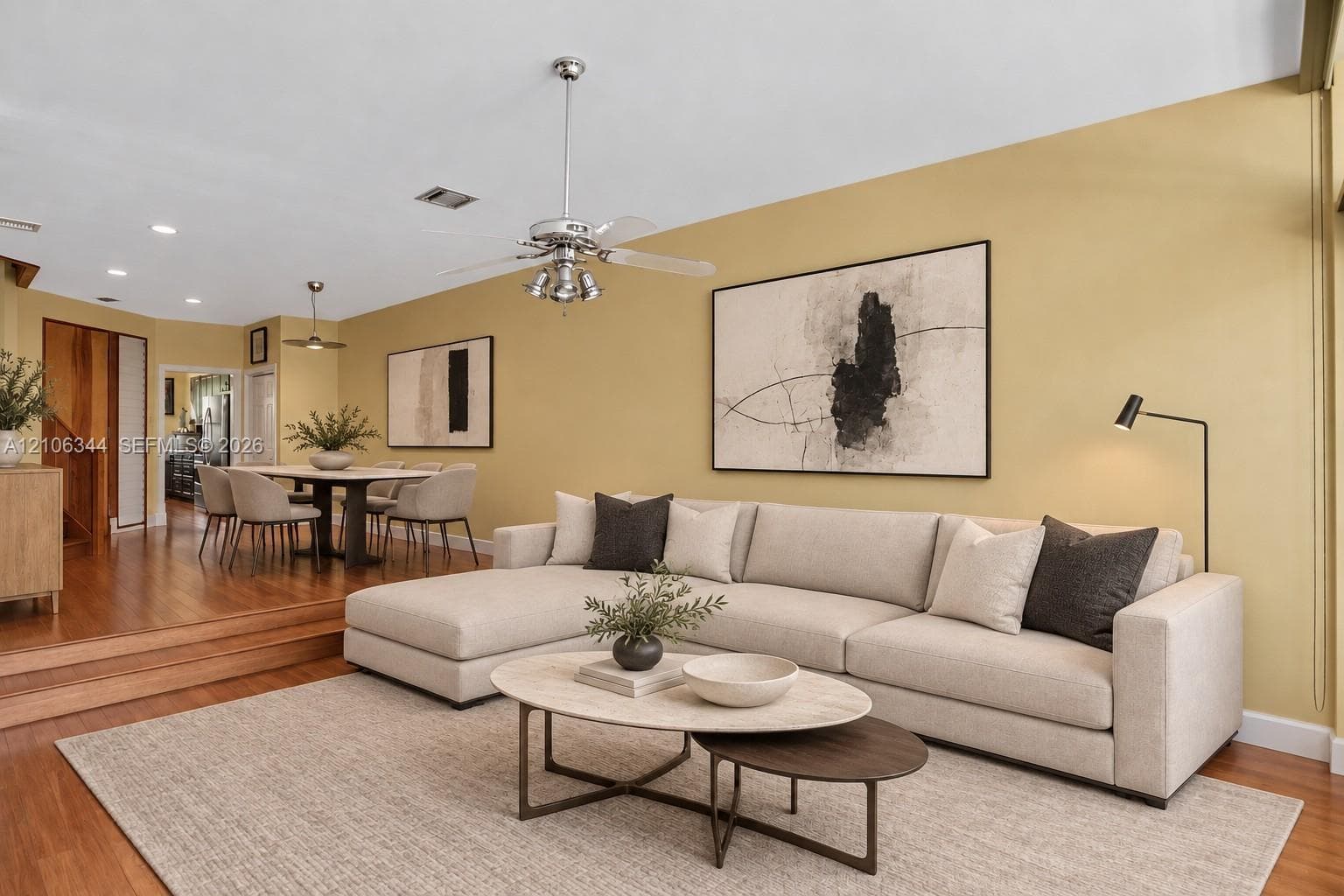 Open-concept living area featuring warm yellow walls, a spacious cream upholstered sectional, wood-finish flooring throughout, recessed lighting with ceiling fan