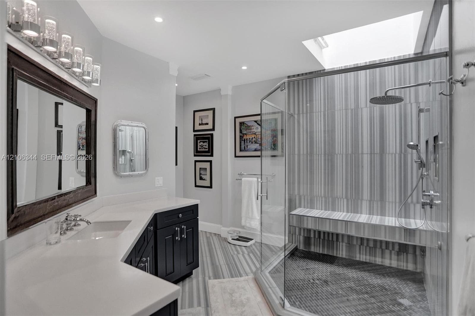 Bathroom featuring a glass-enclosed shower with a built-in bench, dual-sink vanity with a solid-surface countertop, dark cabinetry, and a skylight