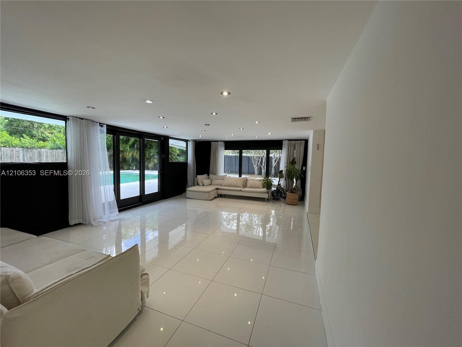 Expansive living area featuring large-format tile flooring, recessed ceiling lighting, and floor-to-ceiling glass sliding doors opening to an outdoor swimming pool