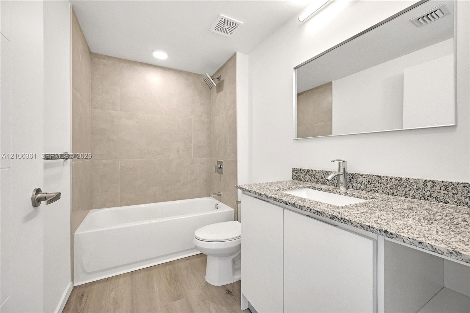 Bathroom featuring a white bathtub, beige tile tub surround, wood-finish flooring, stone-surface vanity countertop with a rectangular undermount sink, and a full-width wall-mounted mirror