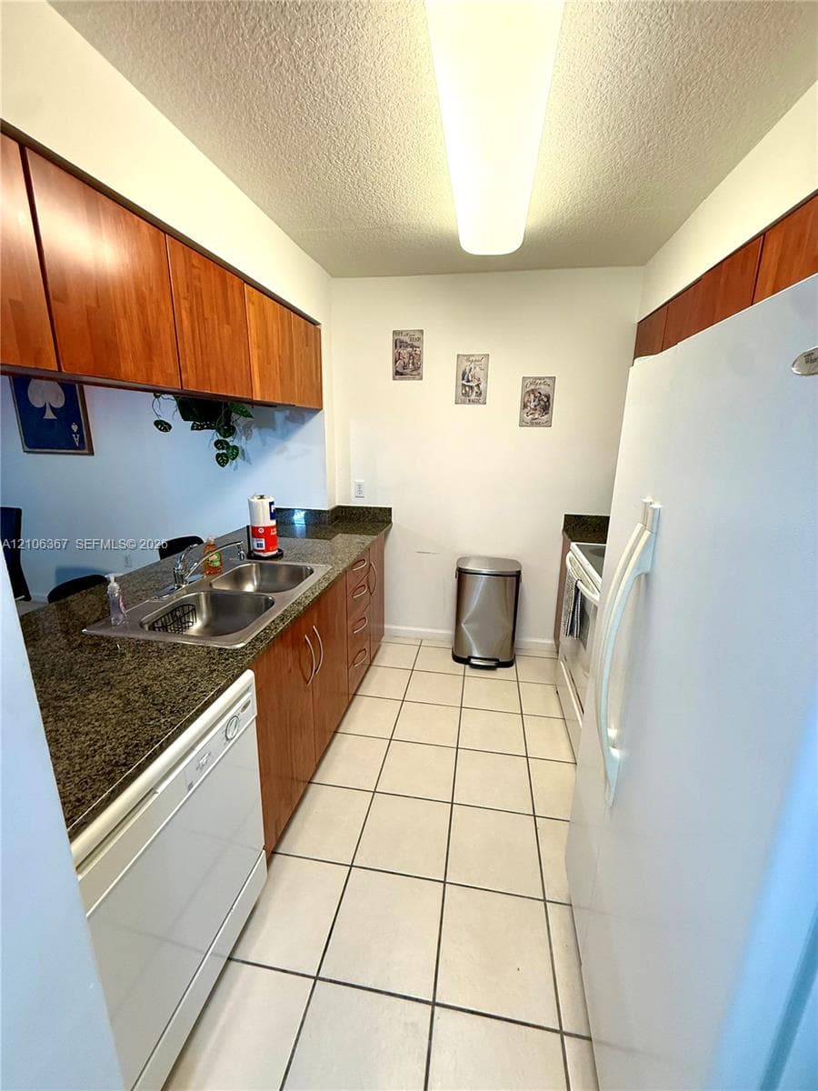 Galley kitchen featuring wood-finish cabinetry, stone-texture countertops, integrated double-basin stainless steel sink, and ceramic tile flooring