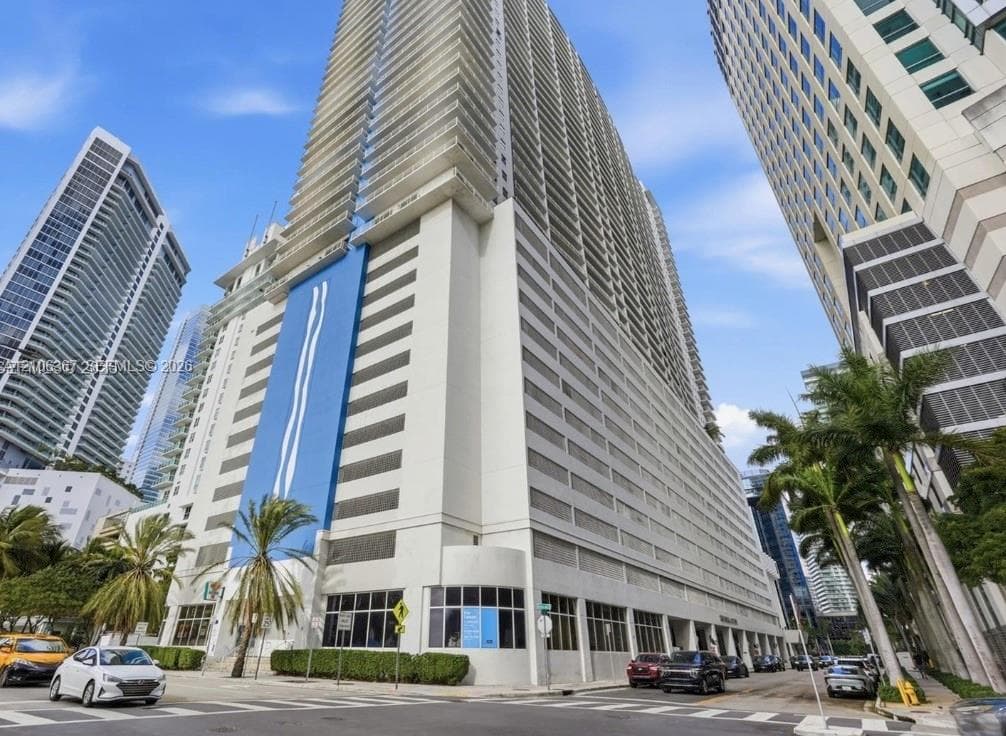 High-rise tower exterior featuring a smooth stucco façade, glass-panel balcony railings, multi-story integrated parking structure, and floor-to-ceiling windows