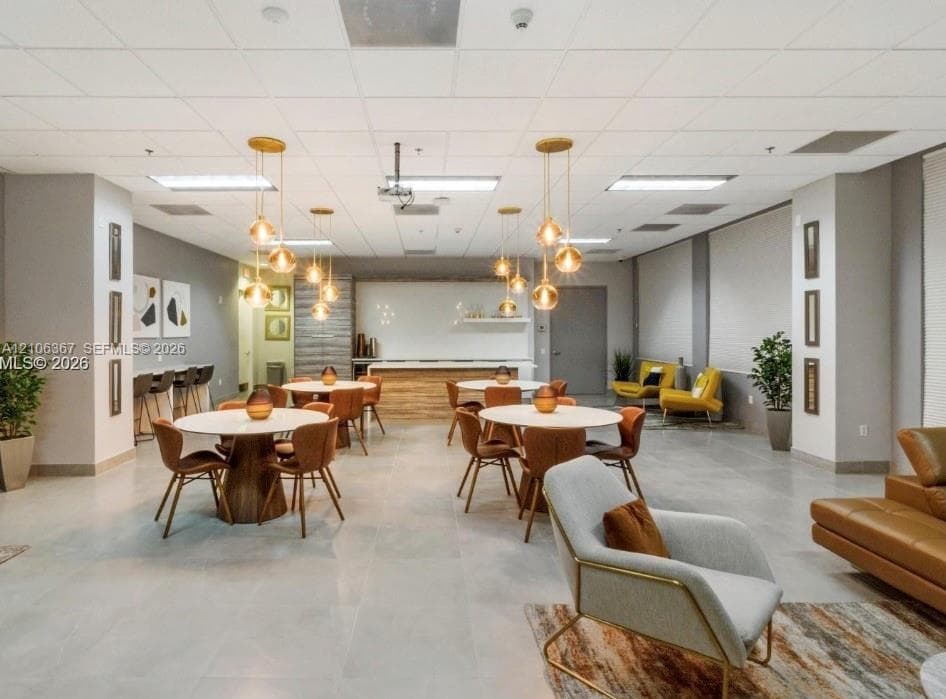 Open-concept common area featuring large-format tile flooring, a built-in refreshment bar with quartz-style countertops, drop ceiling grid, and multiple gold-tone pendant light fixtures