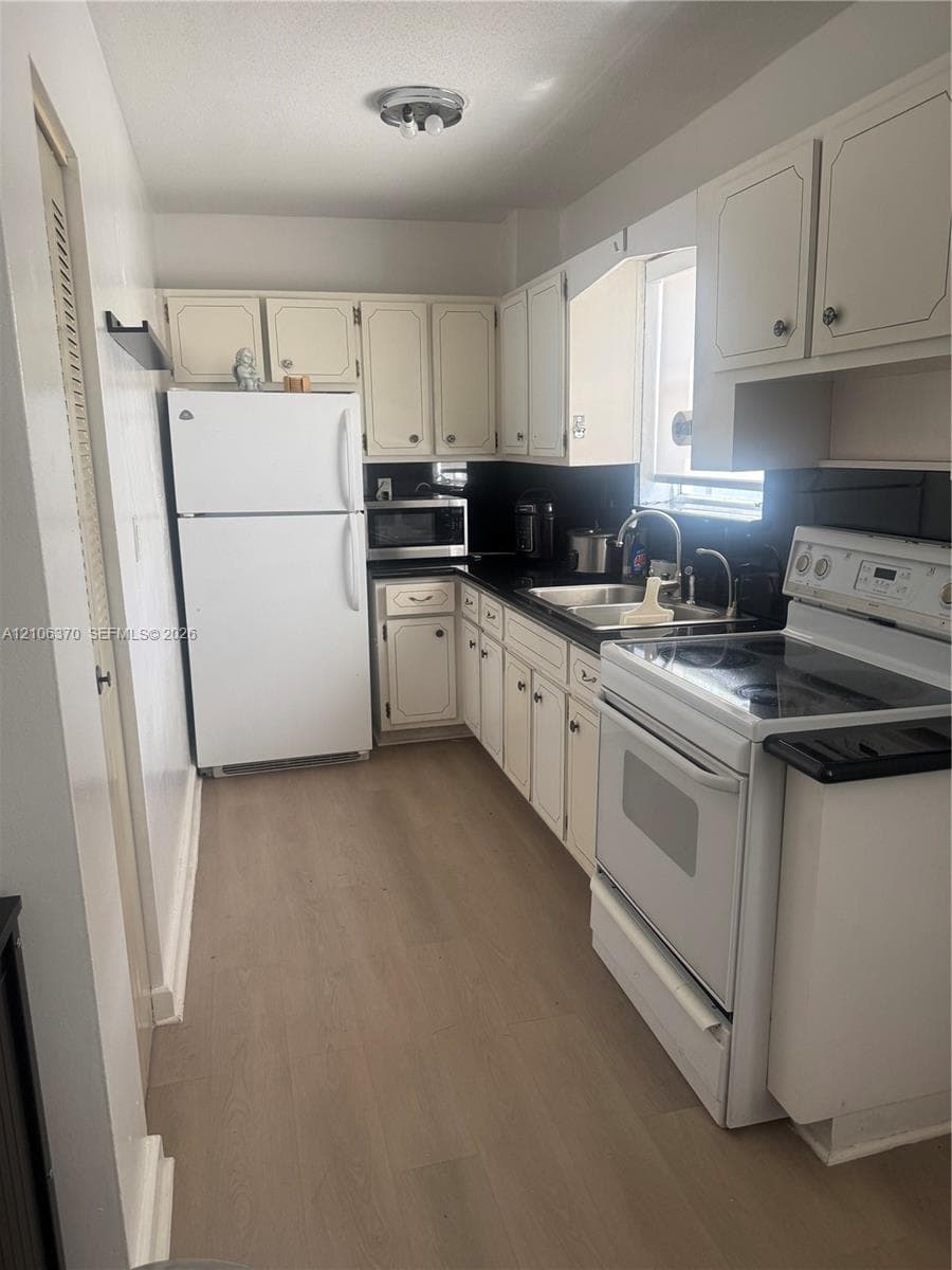 Kitchen featuring white cabinetry, wood-finish flooring, dark solid-surface countertops, dual-basin stainless steel sink, and white appliances