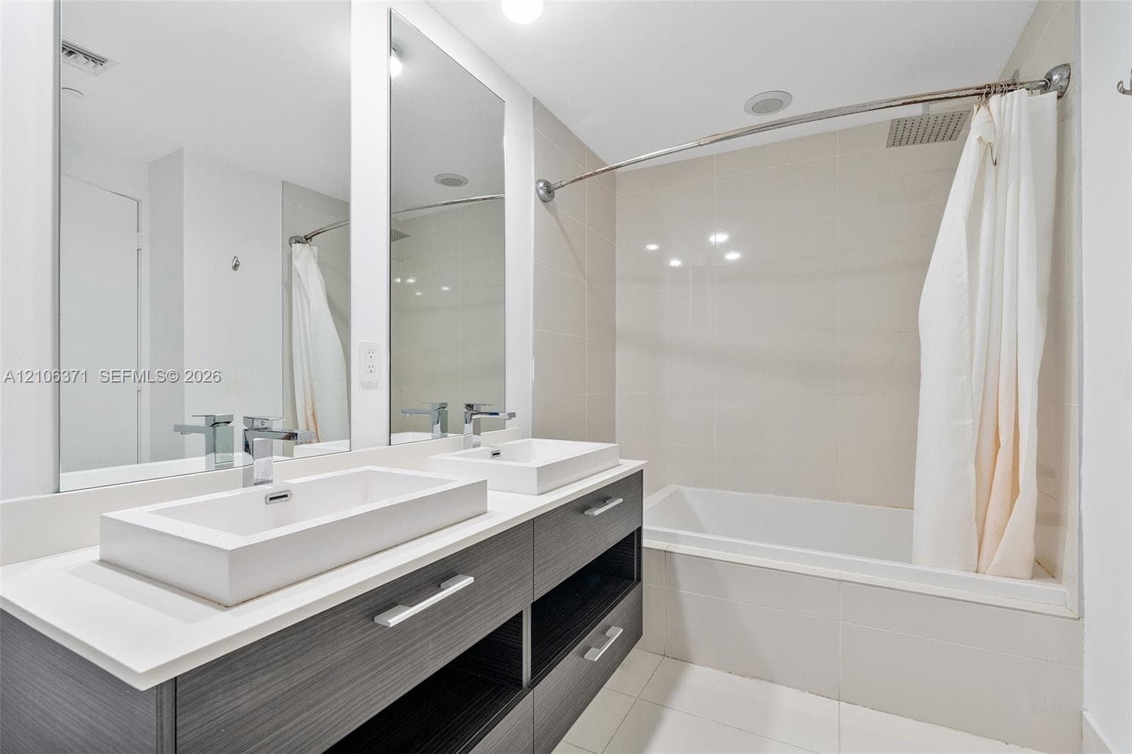 Double vanity featuring a light-toned countertop with rectangular vessel sinks, dark-finish cabinetry, full-width mirror, and a tile-surround bathtub