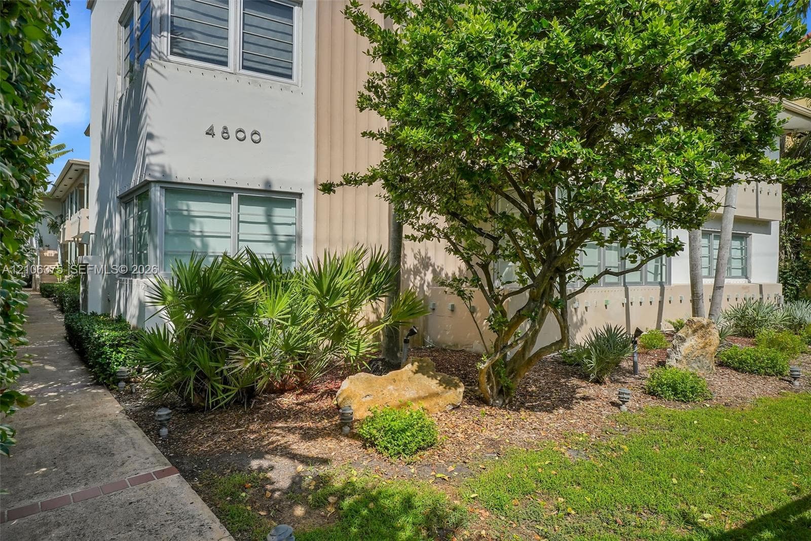 Multi-story exterior featuring a light-toned facade, horizontal casement windows, concrete walkway, and native landscaping with mature trees