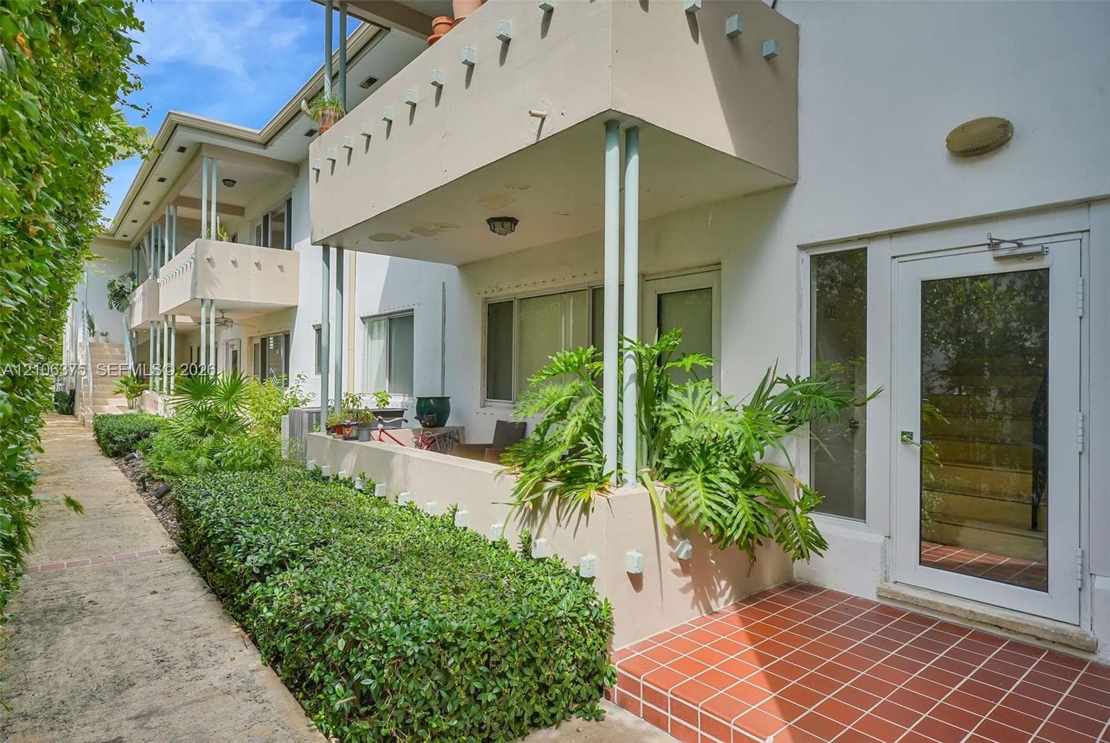 Exterior facade featuring a concrete balcony with decorative notched trim, tile-surfaced patio, and glass-paneled entry door, complemented by manicured shrubbery and a concrete walkway