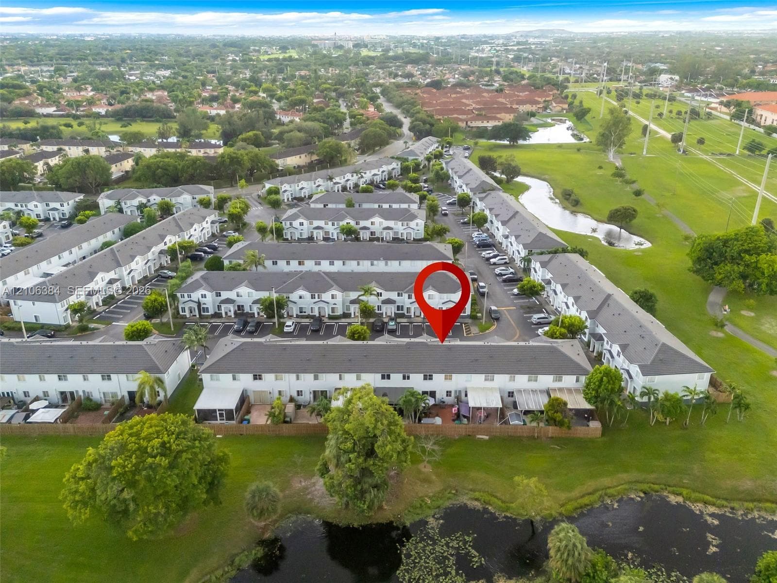 Aerial perspective of multi-family residential architecture featuring pitched shingled roofing, light-toned siding, and paved parking surfaces, situated within a landscape of expansive green space and water features