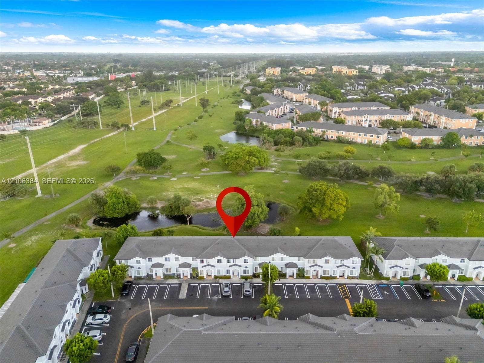 Aerial perspective featuring multi-story residential buildings, paved surface parking, a pond, and expansive green space