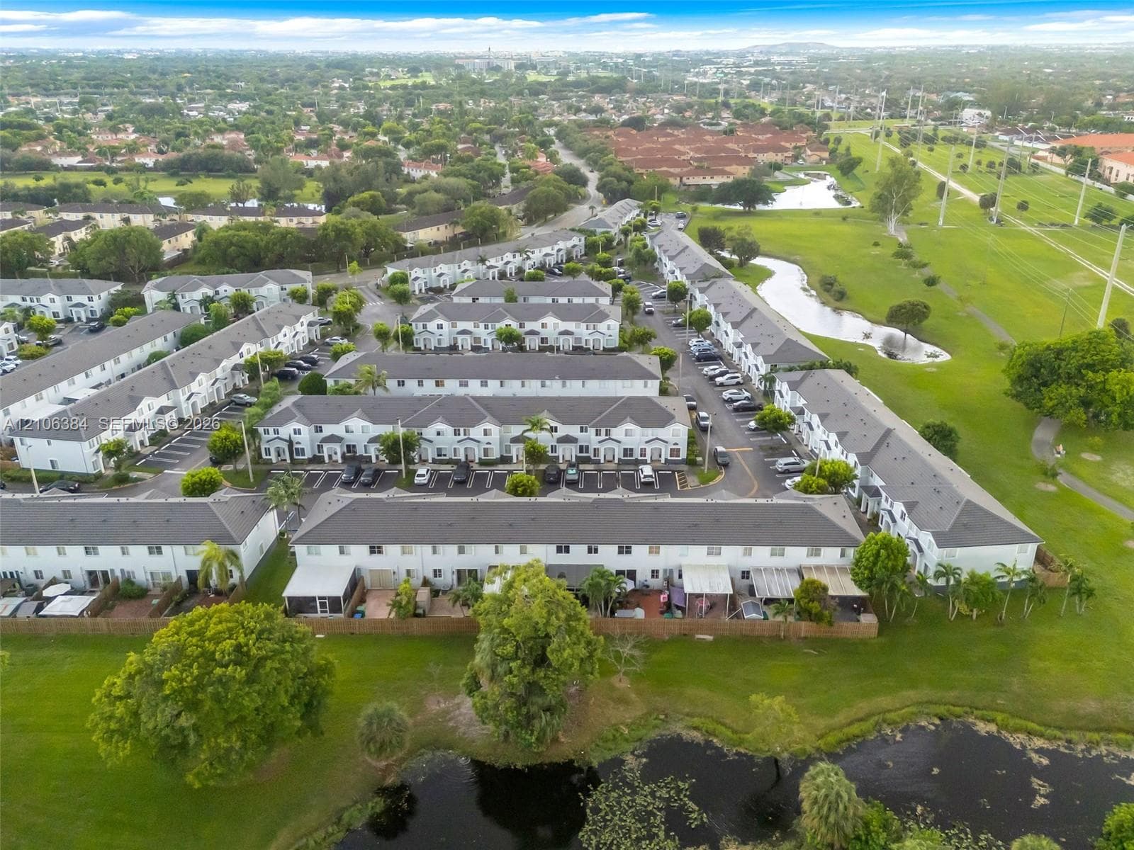 Aerial perspective of multi-family residences featuring grey shingle roofing, exterior light-toned finishes, dedicated parking spaces, and expansive green space with water features