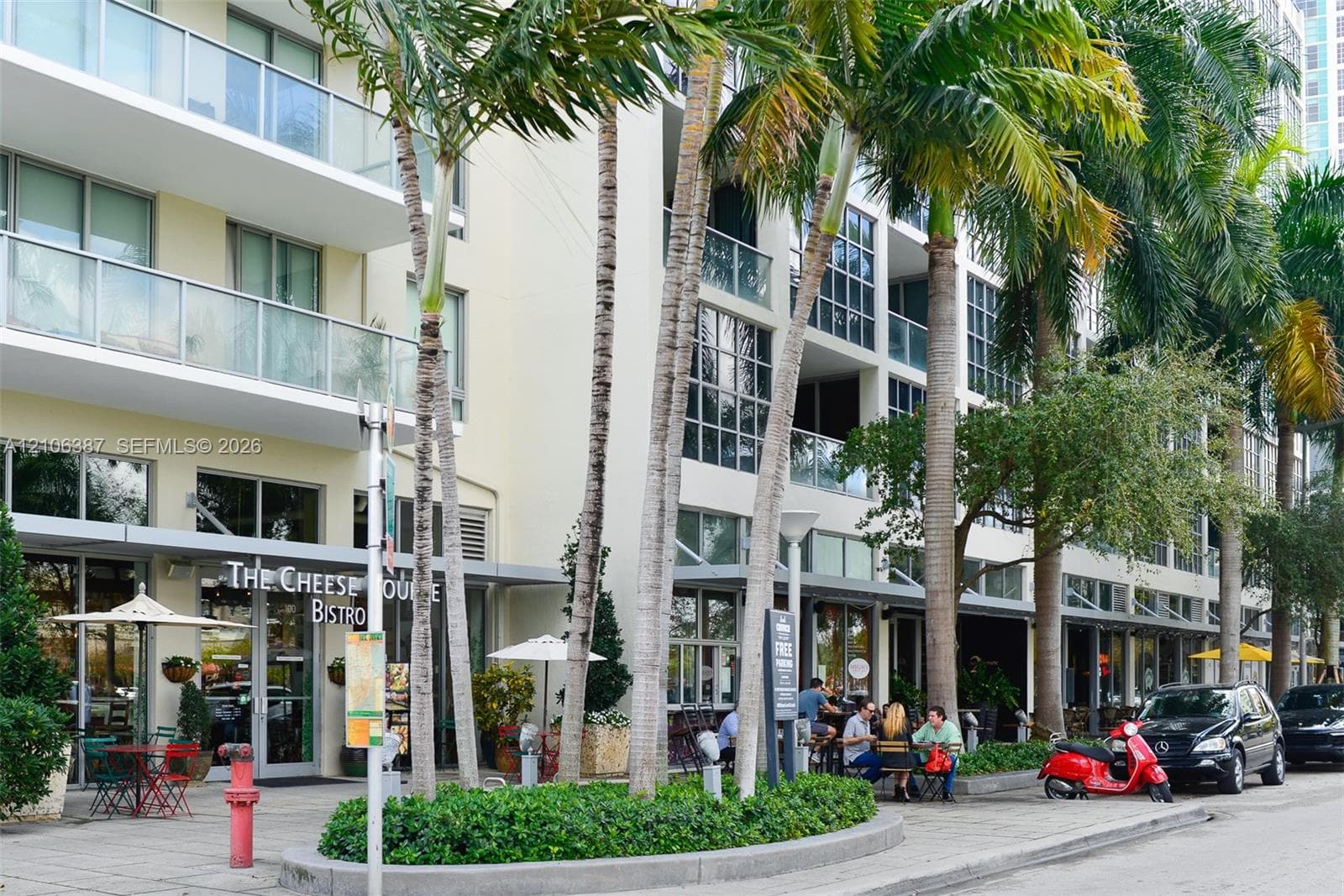 Multi-story building exterior featuring glass-railing balconies, floor-to-ceiling windows, and ground-level commercial storefronts, complemented by mature palm trees and manicured landscape planters
