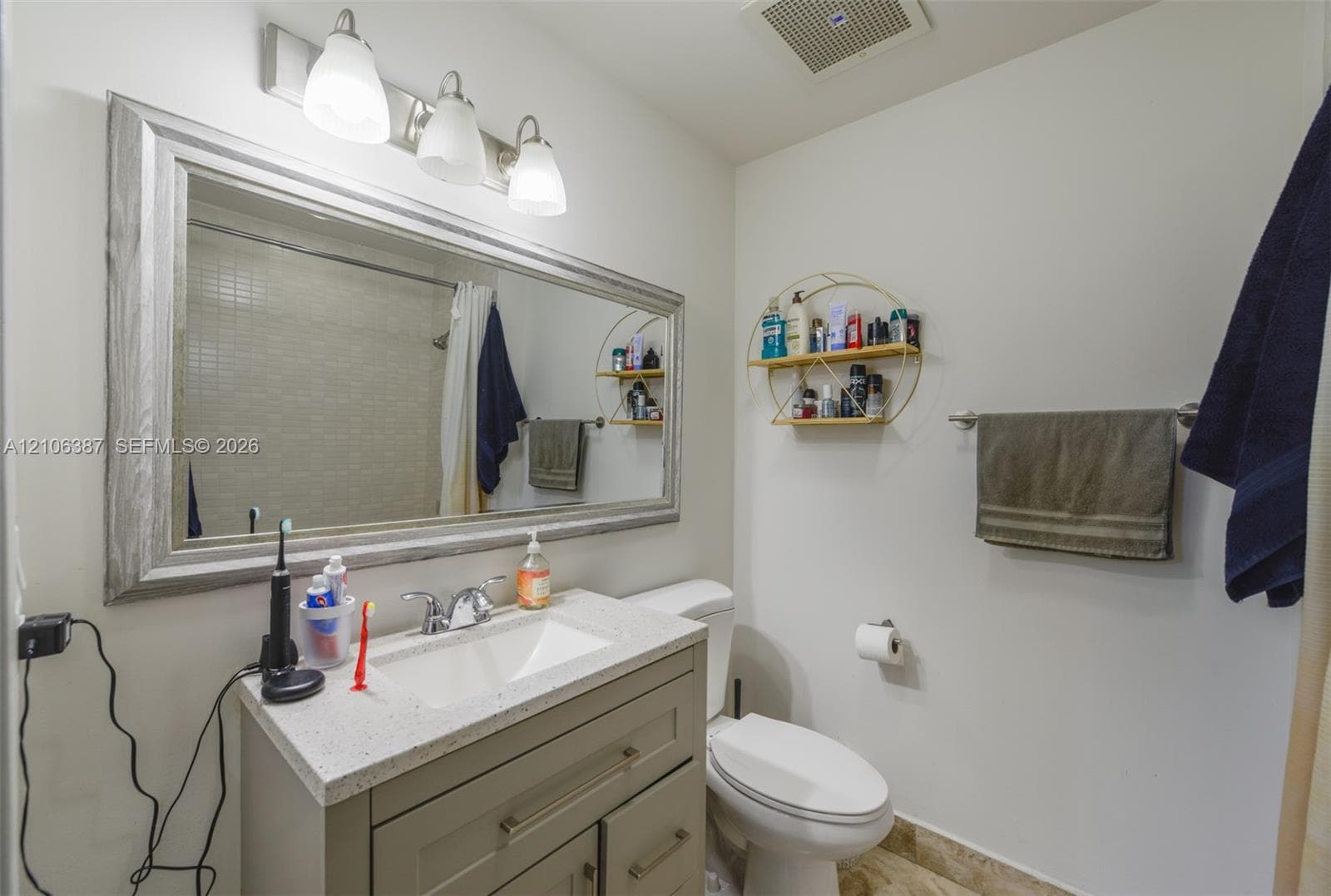 Vanity featuring a stone-composite countertop with an integrated rectangular sink, three-drawer cabinetry with metal hardware, framed wall-mounted mirror, and triple-light vanity fixture