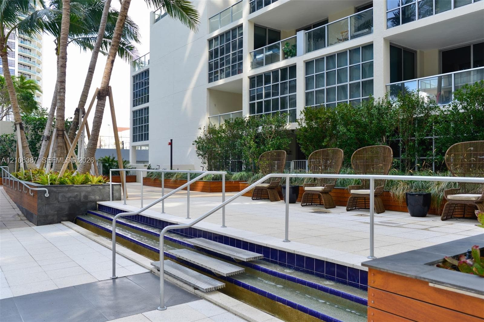 Outdoor terrace featuring tiered tile decking, a linear water feature with blue tile accents, multi-level metal handrails, and integrated planter boxes with wood-finish siding
