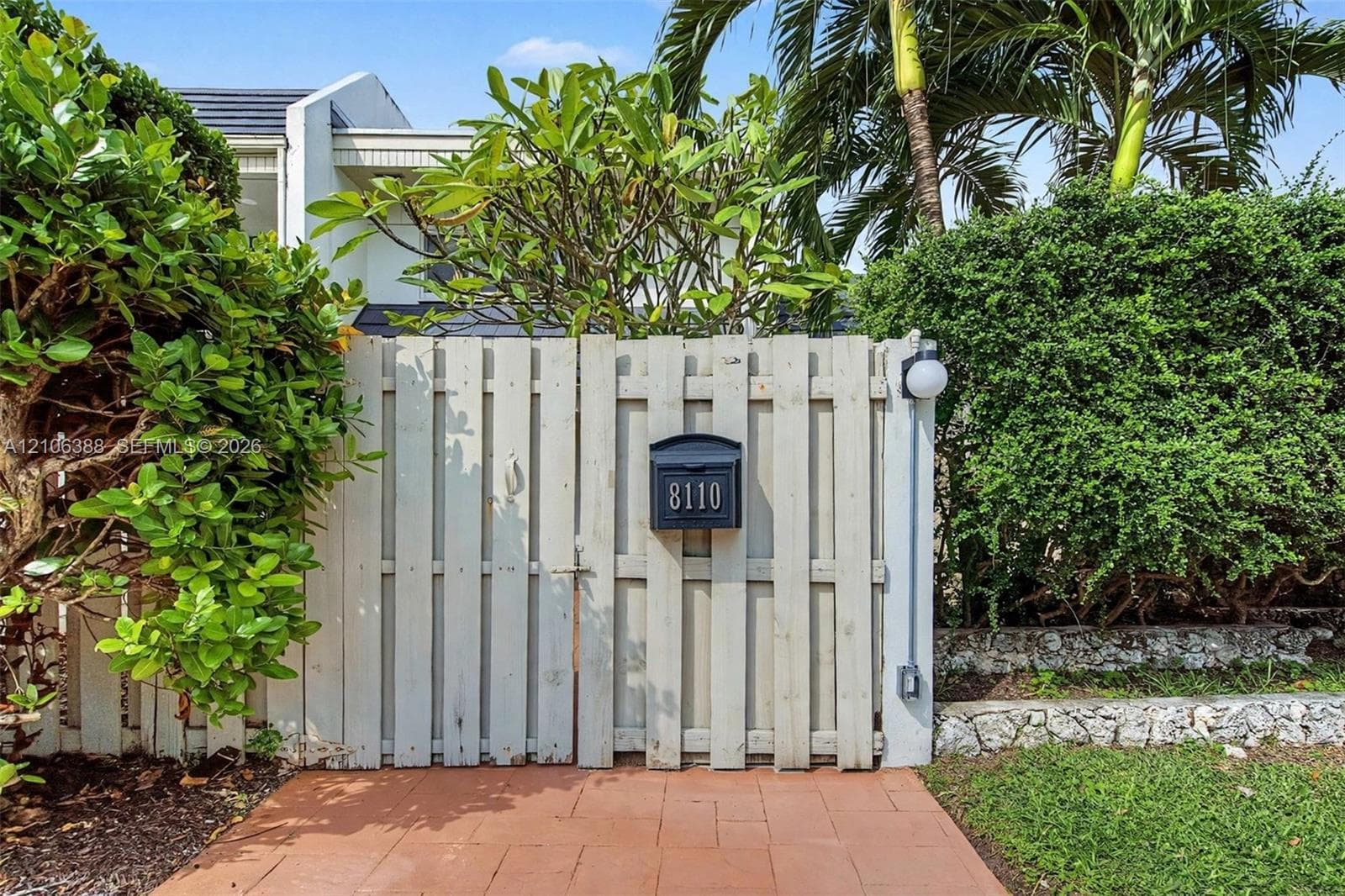 Vertical wood-plank privacy gate featuring a mounted mailbox and globe-style wall sconce, complemented by terracotta-tile paving and natural stone landscaping borders