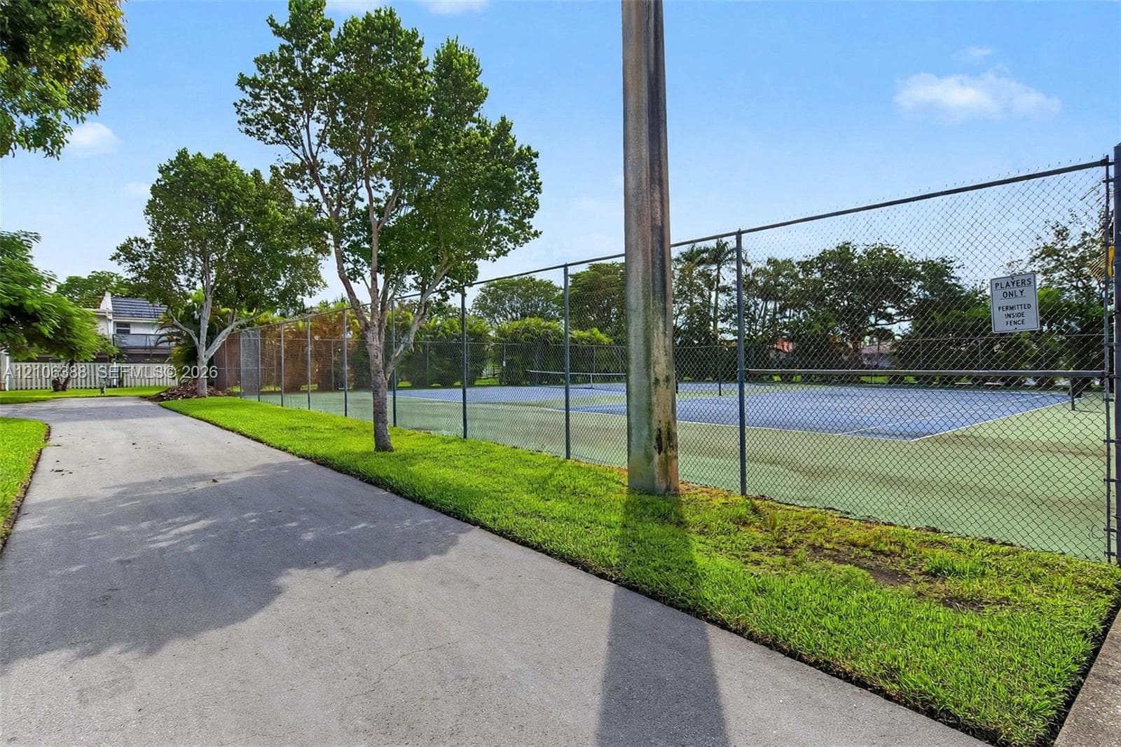 Outdoor recreational area featuring a paved pathway, tennis court with chain-link fencing, and manicured green space