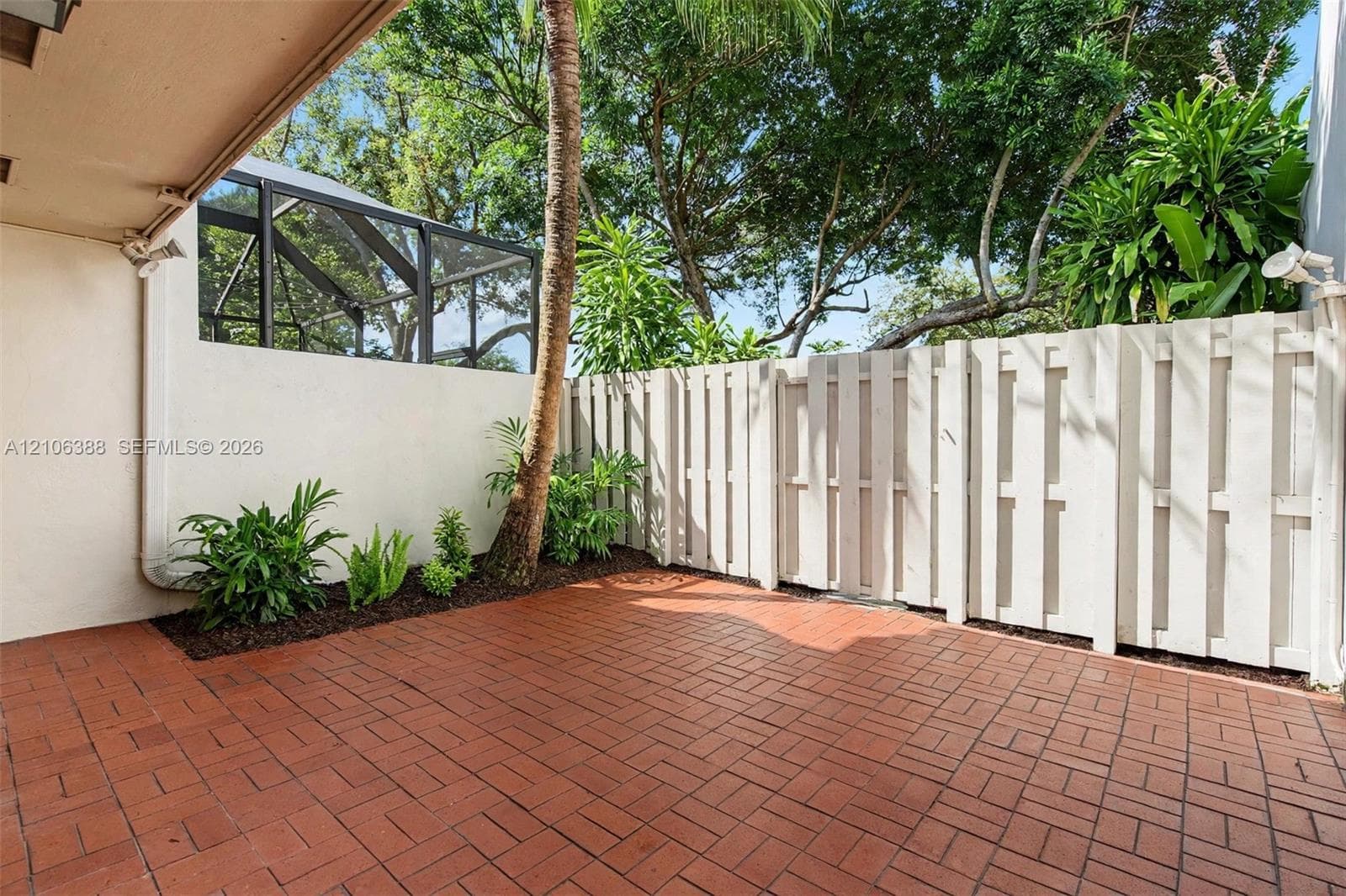 Private patio featuring red brick pavers, a white wood-style privacy fence, and mature landscaping