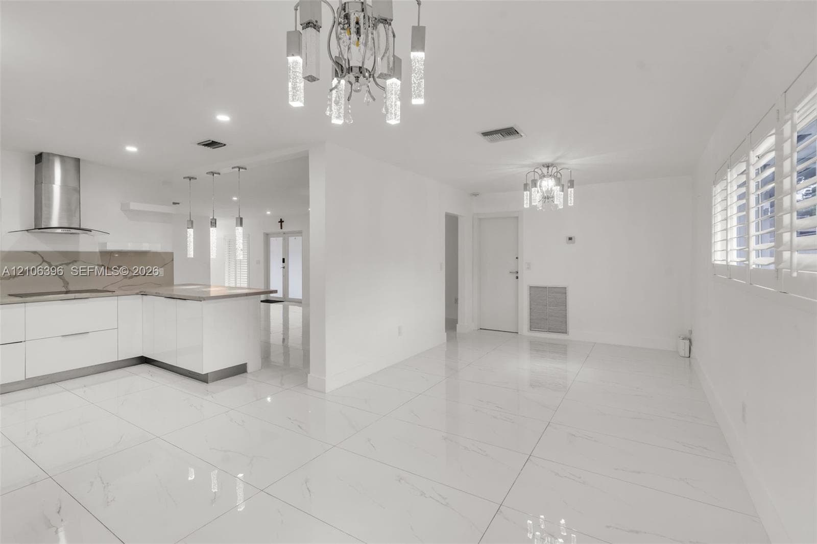 Open-concept interior featuring large-format tile flooring, modern white cabinetry, stone-finish countertops, stainless steel range hood, and crystal-accented chandeliers