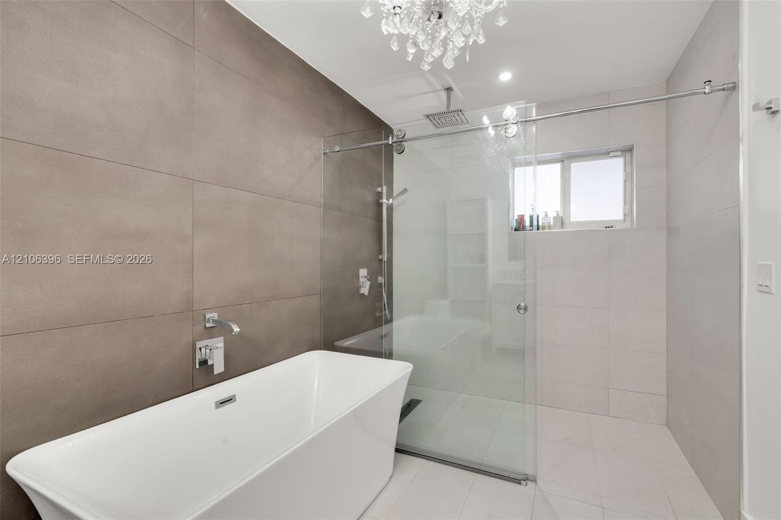 Freestanding soaking tub with wall-mounted polished chrome faucet, glass-enclosed walk-in shower with rainfall showerhead, large-format neutral wall tiling, and crystal chandelier fixture